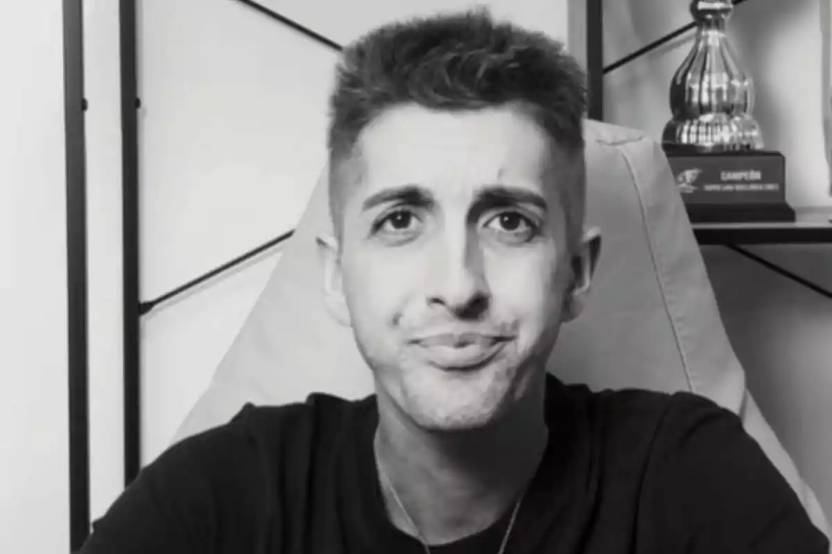 Persona joven con expresión facial de duda o confusión sentada frente a un estante con un trofeo al fondo en una imagen en blanco y negro