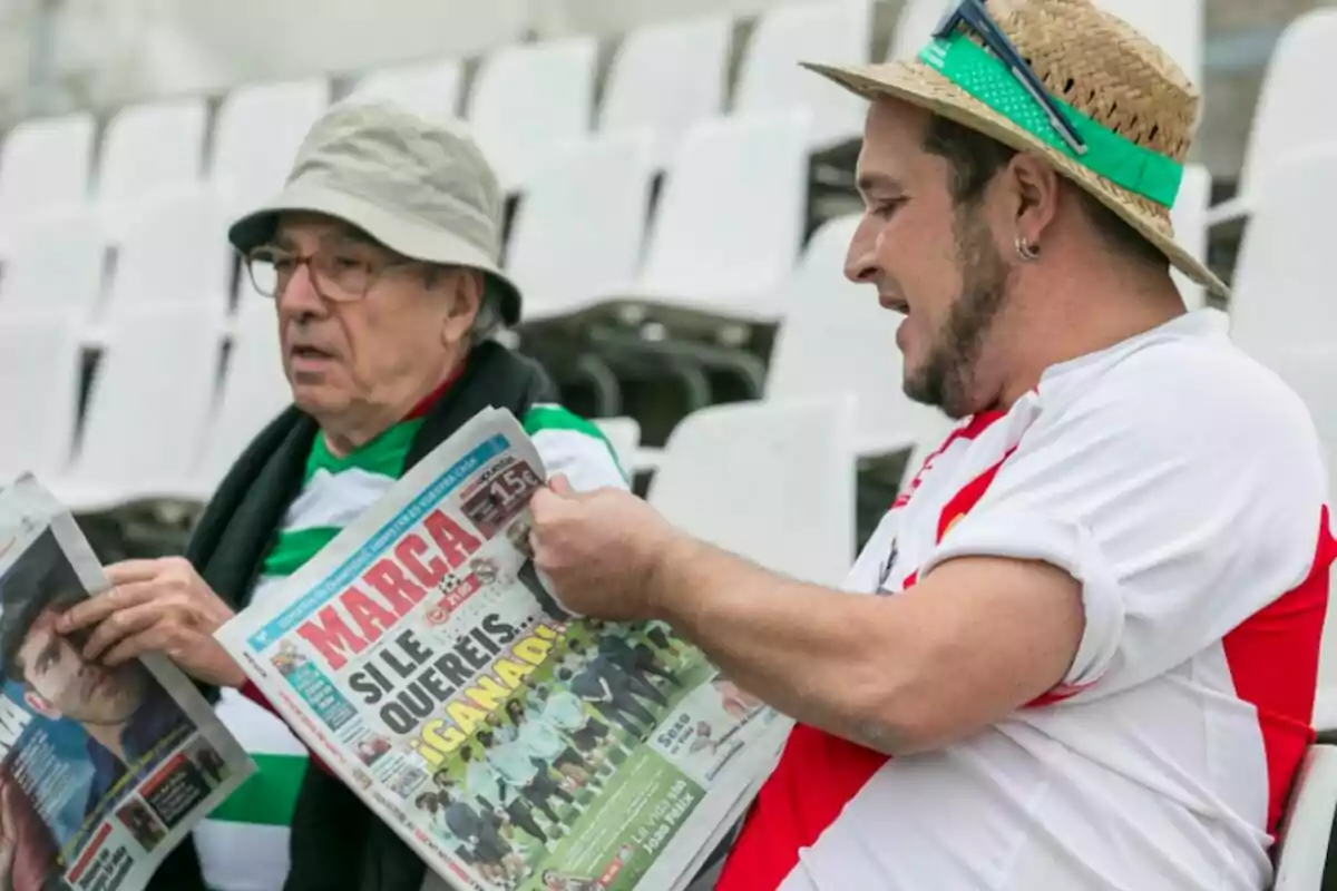Dos hombres sentados en gradas vacías leen periódicos deportivos y conversan animadamente Dos hombres sentados en gradas vacías leen periódicos deportivos y conversan animadamente