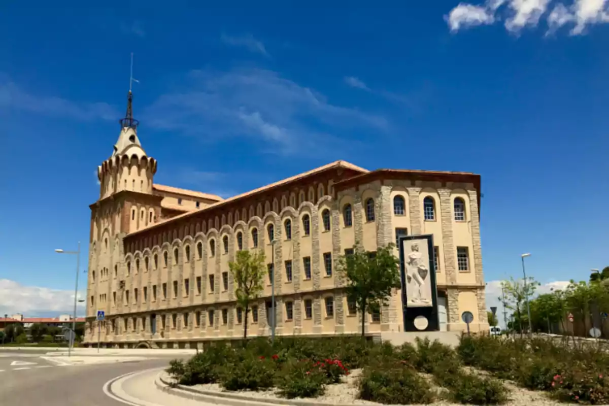 Edificio histórico de varios pisos con una torre en la esquina y una escultura en la fachada rodeado de árboles y arbustos bajo un cielo azul despejado Edificio histórico de varios pisos con una torre en la esquina y una escultura en la fachada rodeado de árboles y arbustos bajo un cielo azul despejado