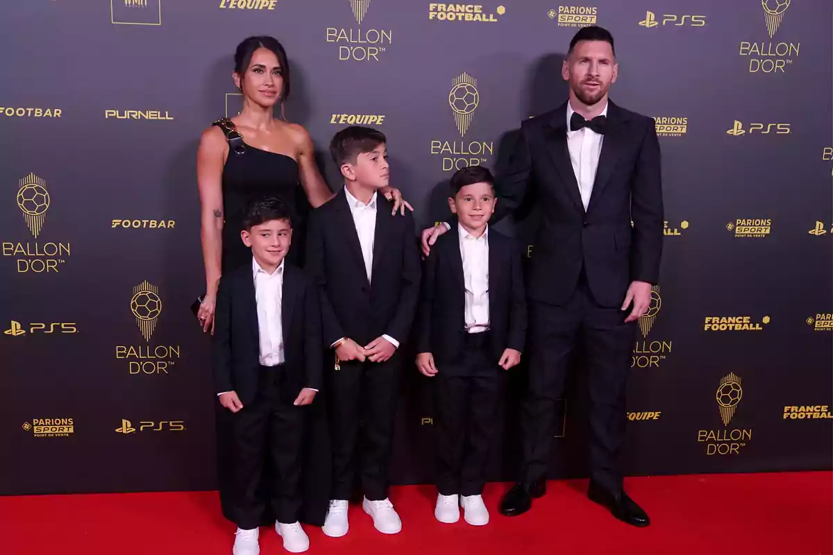 An elegantly dressed family poses on the red carpet at the Ballon d'Or ceremony, with a black background decorated with golden event logos. An elegantly dressed family poses on the red carpet at the Ballon d'Or ceremony, with a black background decorated with golden event logos.