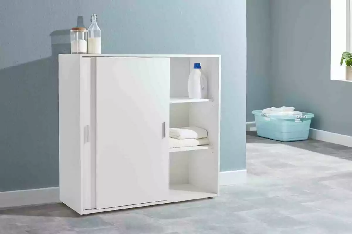 White cabinet with sliding door in a room, with bottles and a jar on top, towels and detergent on the shelves, and a blue basket in the back. White cabinet with sliding door in a room, with bottles and a jar on top, towels and detergent on the shelves, and a blue basket in the back.