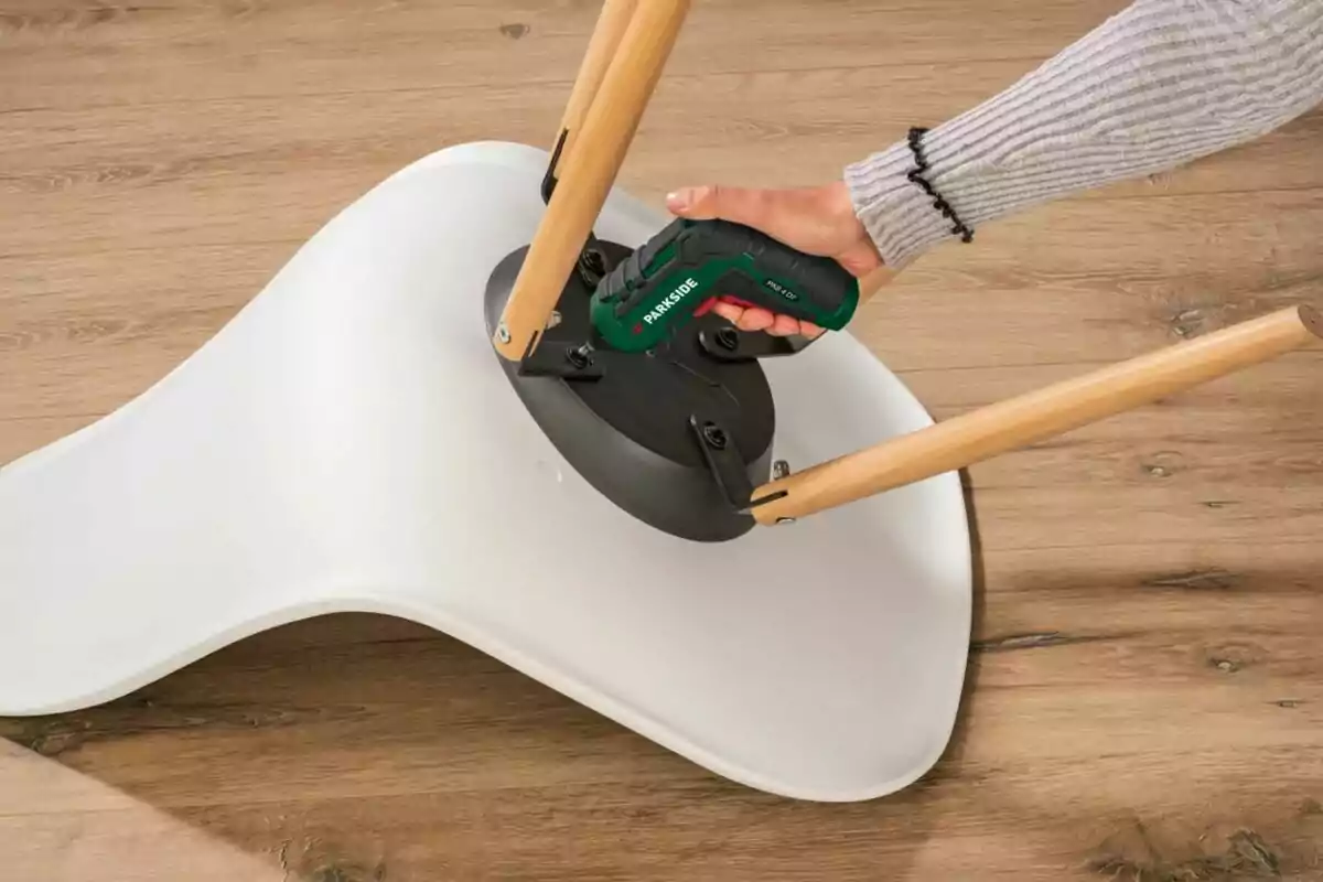 Persona usando un destornillador eléctrico para ensamblar las patas de una silla blanca sobre un suelo de madera Persona usando un destornillador eléctrico para ensamblar las patas de una silla blanca sobre un suelo de madera