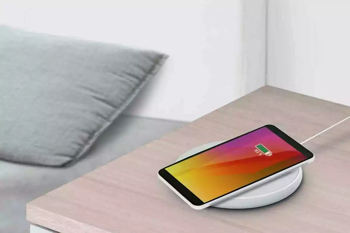 Un teléfono móvil cargándose de manera inalámbrica sobre una mesa junto a un cojín gris Un teléfono móvil cargándose de manera inalámbrica sobre una mesa junto a un cojín gris