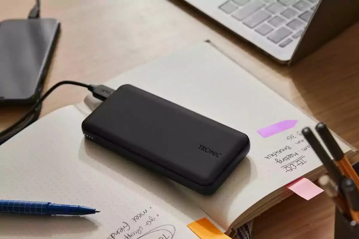 Bateria portàtil negra sobre una llibreta al costat d’un bolígraf blau, un telèfon mòbil i un ordinador portàtil en un escriptori de fusta Bateria portàtil negra sobre una llibreta al costat d’un bolígraf blau, un telèfon mòbil i un ordinador portàtil en un escriptori de fusta