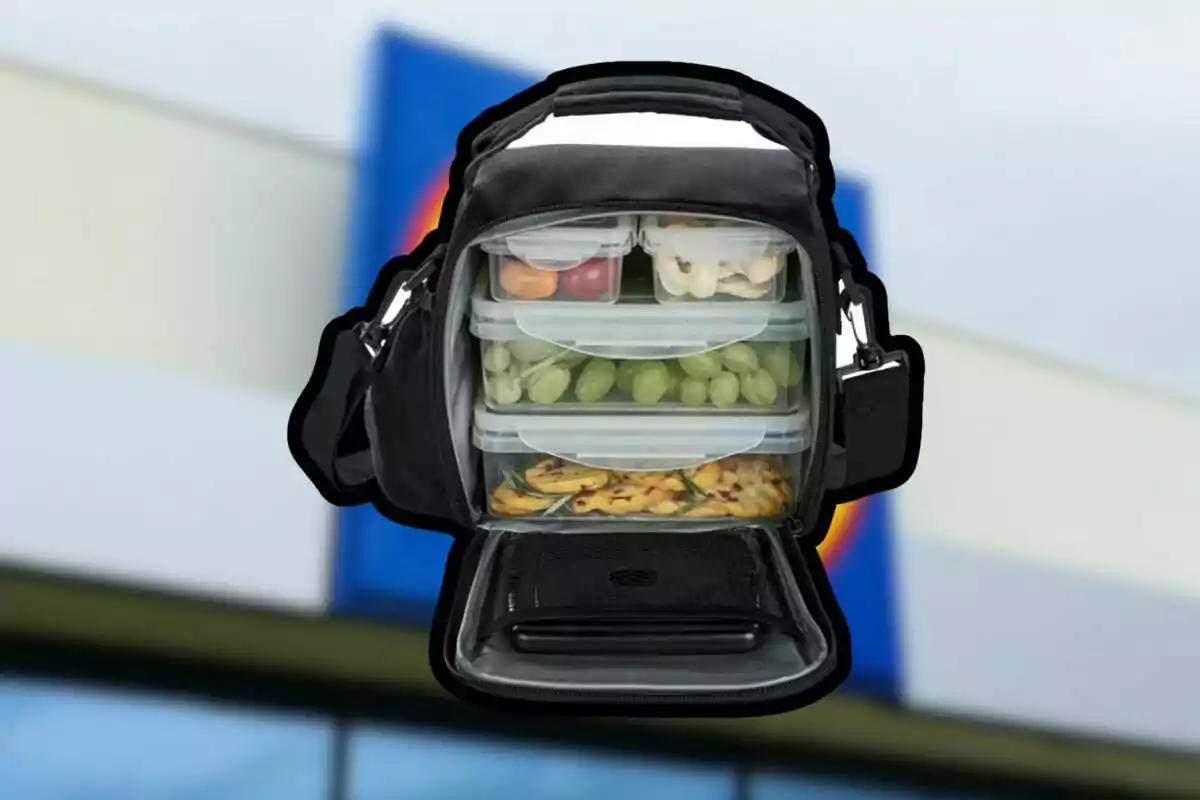 Bolsa térmica negra abierta mostrando varios recipientes transparentes con comida como uvas, tomates, pan y snacks frente a un fondo borroso de edificio Bolsa térmica negra abierta mostrando varios recipientes transparentes con comida como uvas, tomates, pan y snacks frente a un fondo borroso de edificio