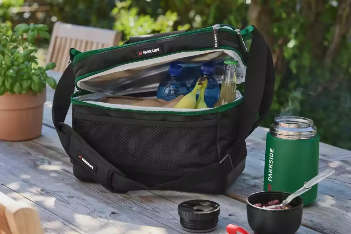 Black thermal bag with a green zipper on a wooden outdoor table, containing water bottles, bananas, and a bottle, next to a green thermos and a food container Black thermal bag with a green zipper on a wooden outdoor table, containing water bottles, bananas, and a bottle, next to a green thermos and a food container