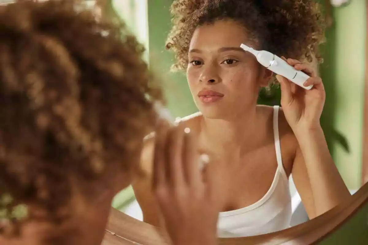 Woman grooming her eyebrows with an electric device in front of a mirror Woman grooming her eyebrows with an electric device in front of a mirror