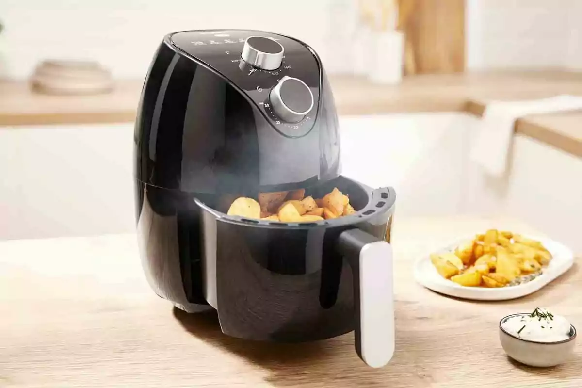 Fregidora d’aire negra cuinant patates en una cuina moderna amb plats de patates i salsa al fons Fregidora d’aire negra cuinant patates en una cuina moderna amb plats de patates i salsa al fons