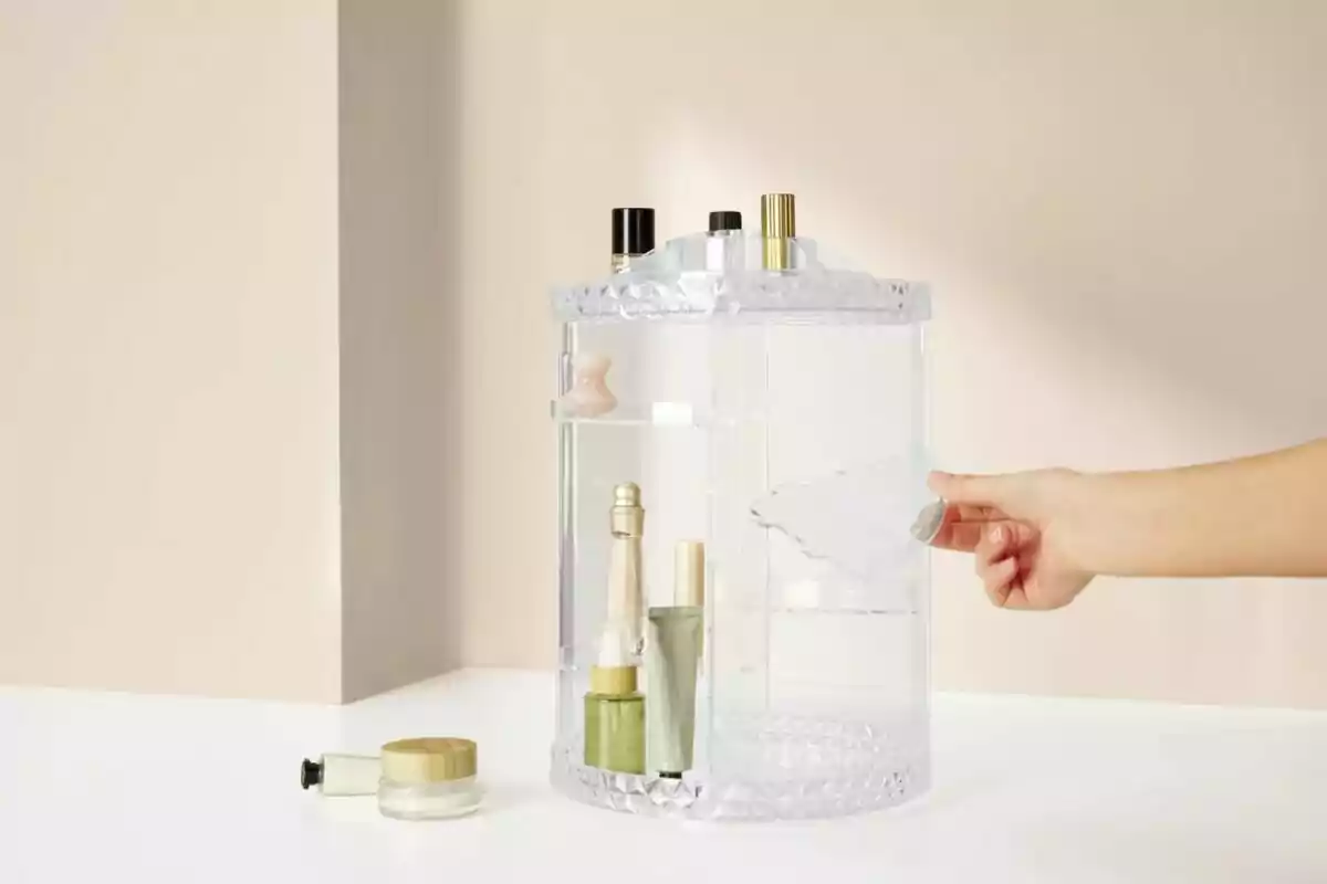 A hand holds the door of a transparent cosmetics organizer that contains several bottles and containers on a white surface. A hand holds the door of a transparent cosmetics organizer that contains several bottles and containers on a white surface.