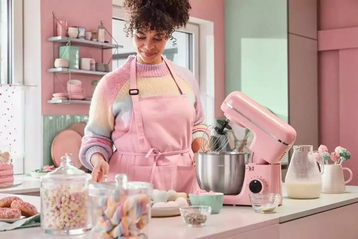 Mujer con delantal rosa preparando repostería en una cocina decorada en tonos pastel con una batidora rosa y frascos de dulces sobre la mesa Mujer con delantal rosa preparando repostería en una cocina decorada en tonos pastel con una batidora rosa y frascos de dulces sobre la mesa