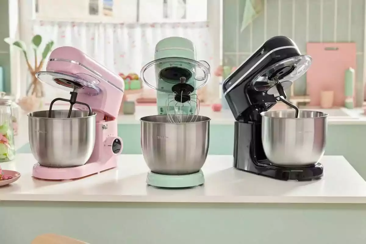 Tres batidoras de pie en colores rosa, verde menta y negro están alineadas sobre una encimera en una cocina iluminada Tres batidoras de pie en colores rosa, verde menta y negro están alineadas sobre una encimera en una cocina iluminada