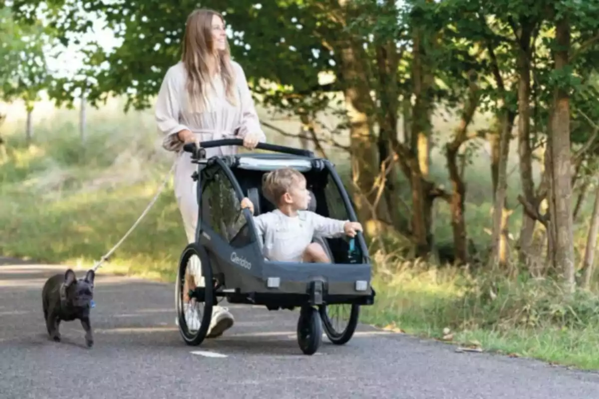 Mujer paseando a un niño en un carrito y llevando a un perro por un camino rodeado de árboles Mujer paseando a un niño en un carrito y llevando a un perro por un camino rodeado de árboles