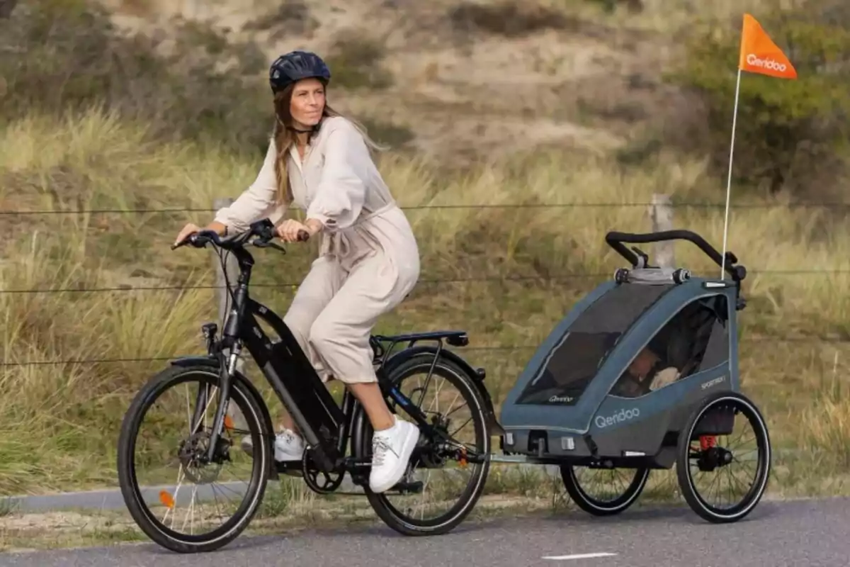 Mujer andando en bicicleta eléctrica con casco y remolque para niños en un camino junto a vegetación Mujer andando en bicicleta eléctrica con casco y remolque para niños en un camino junto a vegetación