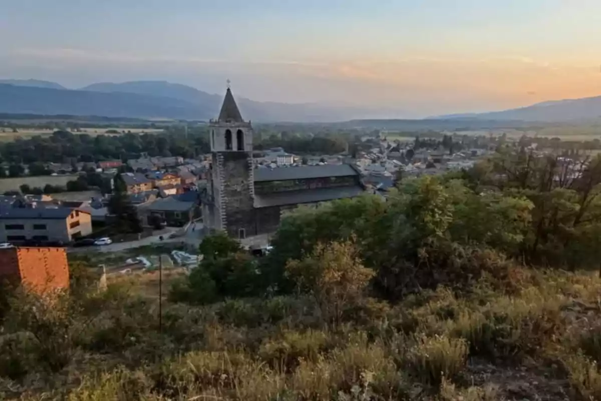 Vista panorámica de un pueblo con una iglesia de torre alta rodeada de casas, vegetación y montañas al fondo al atardecer Vista panorámica de un pueblo con una iglesia de torre alta rodeada de casas, vegetación y montañas al fondo al atardecer