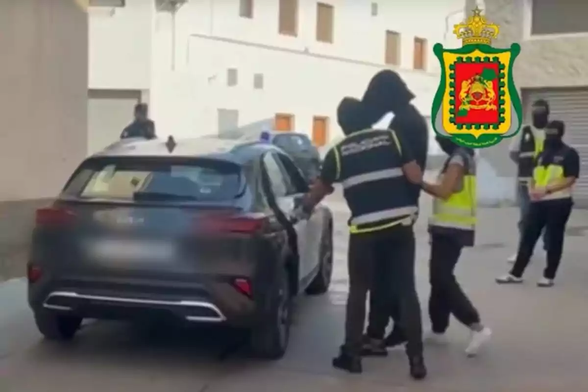 Diversos agents escorten una persona emmascarada cap a un cotxe en un carrer, mentre d’altres observen i un escut oficial apareix a la imatge.