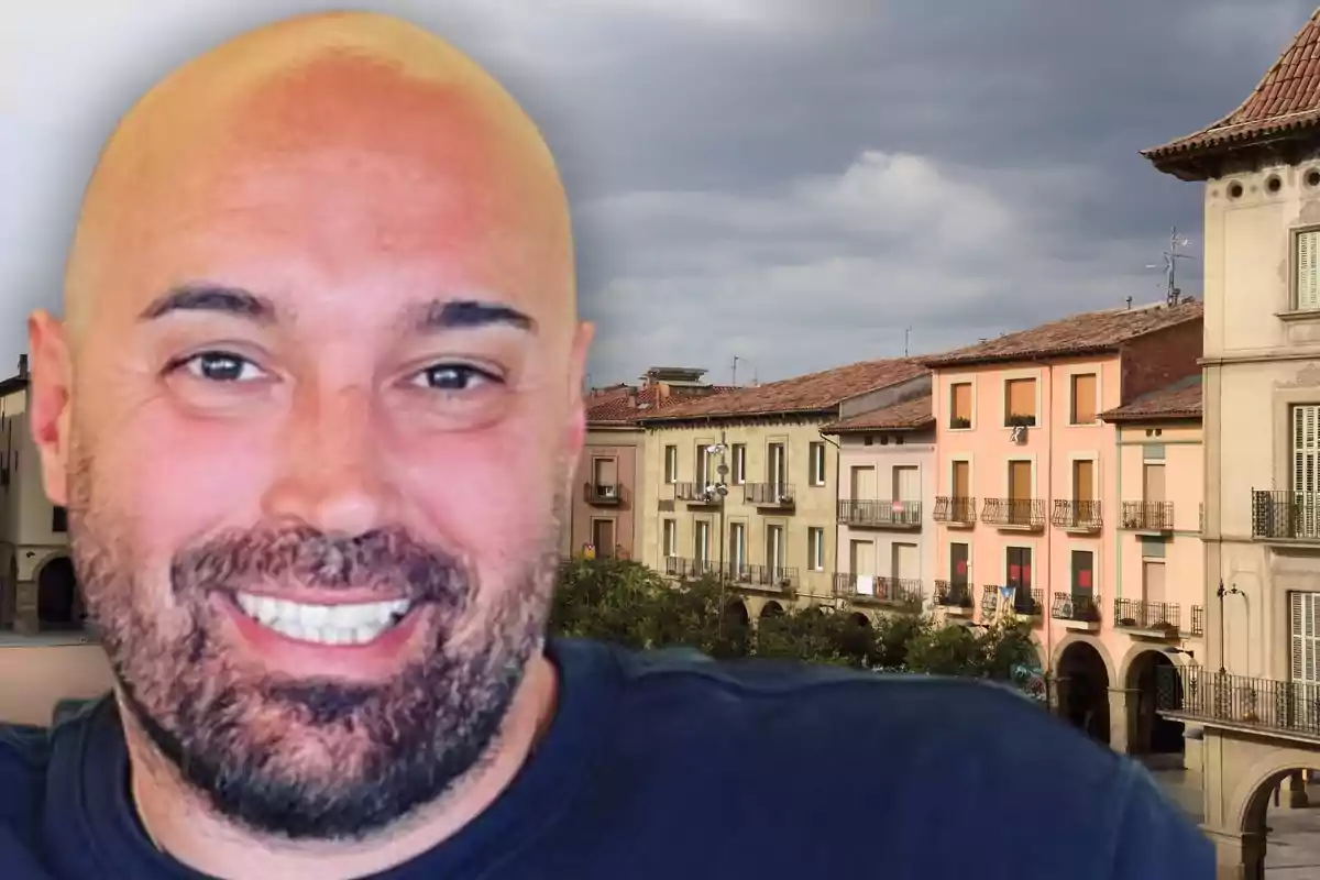 Hombre calvo y sonriente en primer plano con fondo de edificios coloridos y cielo nublado