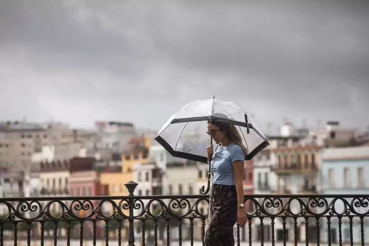Dona caminant sota la pluja amb un paraigua transparent i edificis de colors de fons