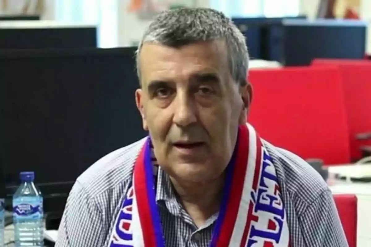 Manuel Esteban Fernández con bufanda de colores rojo, blanco y azul sentado en una oficina con escritorios y una botella de agua a su lado. Manuel Esteban Fernández con bufanda de colores rojo, blanco y azul sentado en una oficina con escritorios y una botella de agua a su lado.