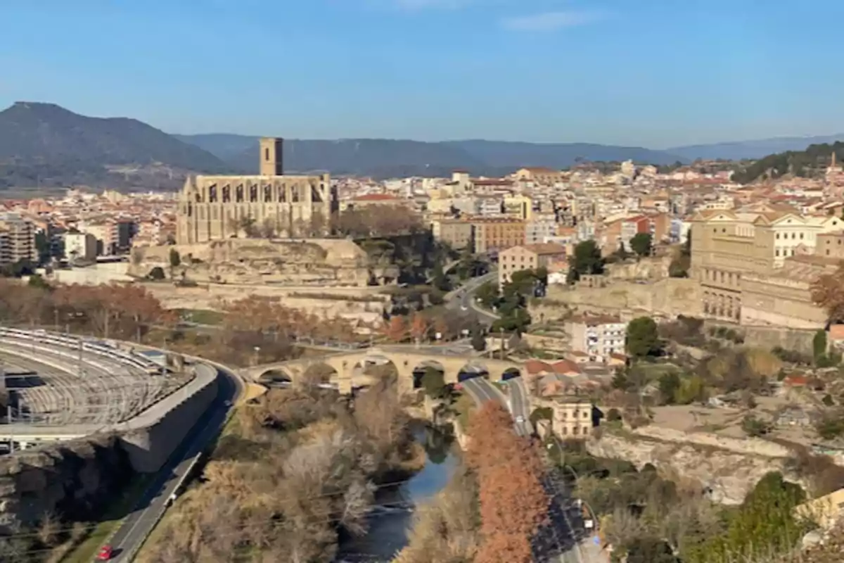 Vista panoràmica d’una ciutat amb una catedral antiga al centre, envoltada d’edificis, muntanyes al fons i un riu que travessa per un pont de pedra Vista panoràmica d’una ciutat amb una catedral antiga al centre, envoltada d’edificis, muntanyes al fons i un riu que travessa per un pont de pedra