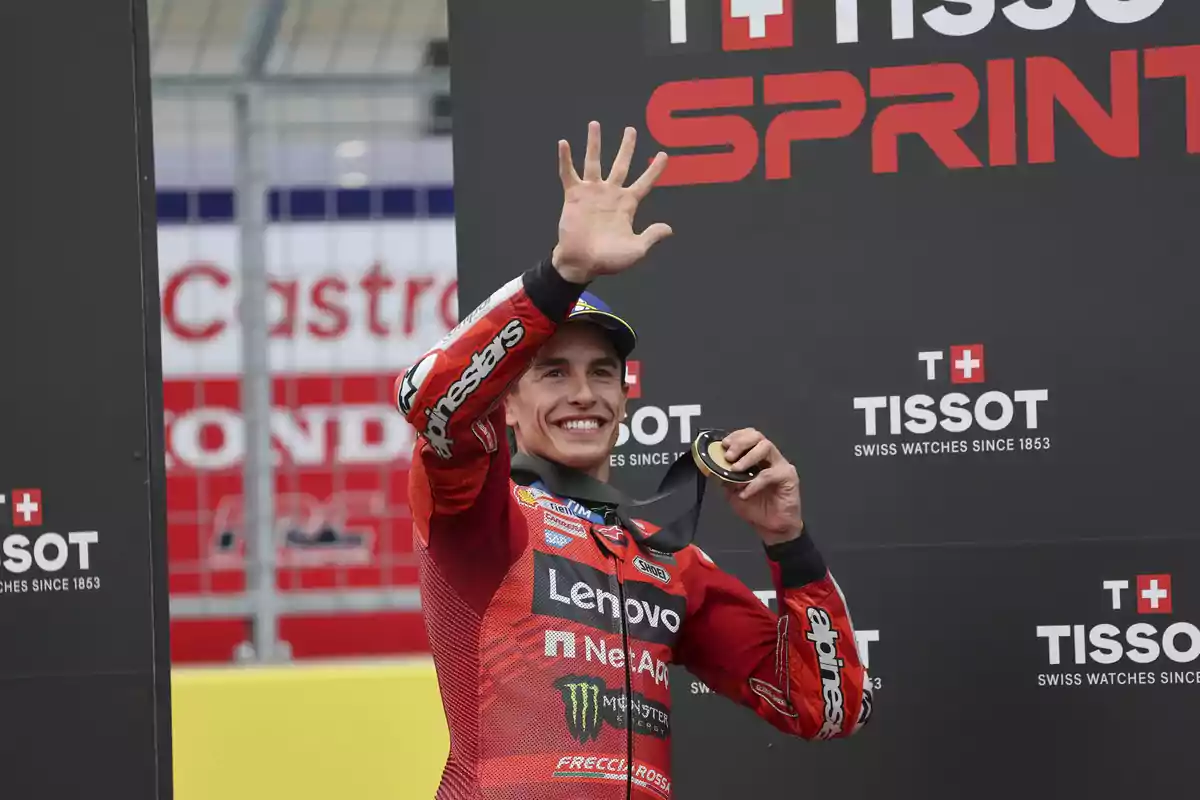 Piloto de motociclismo sonriente con traje rojo celebrando en el podio y mostrando una medalla frente a un fondo con el logo de Tissot Sprint Piloto de motociclismo sonriente con traje rojo celebrando en el podio y mostrando una medalla frente a un fondo con el logo de Tissot Sprint