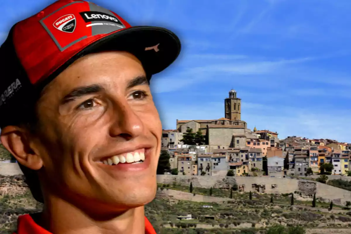 Hombre joven con gorra roja sonriente en primer plano con un pueblo antiguo y una iglesia al fondo bajo un cielo azul