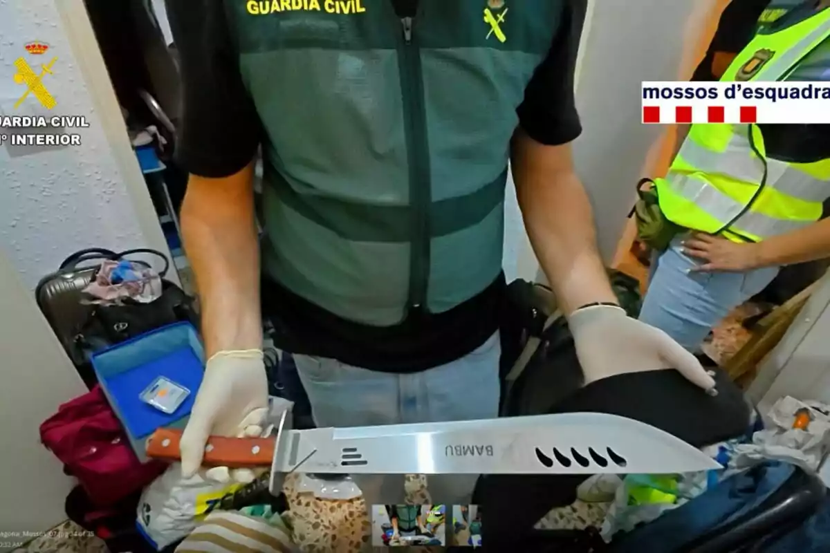 Agent de la Guàrdia Civil sosté un matxet gran amb mànec de fusta durant una operació policial en una habitació desordenada