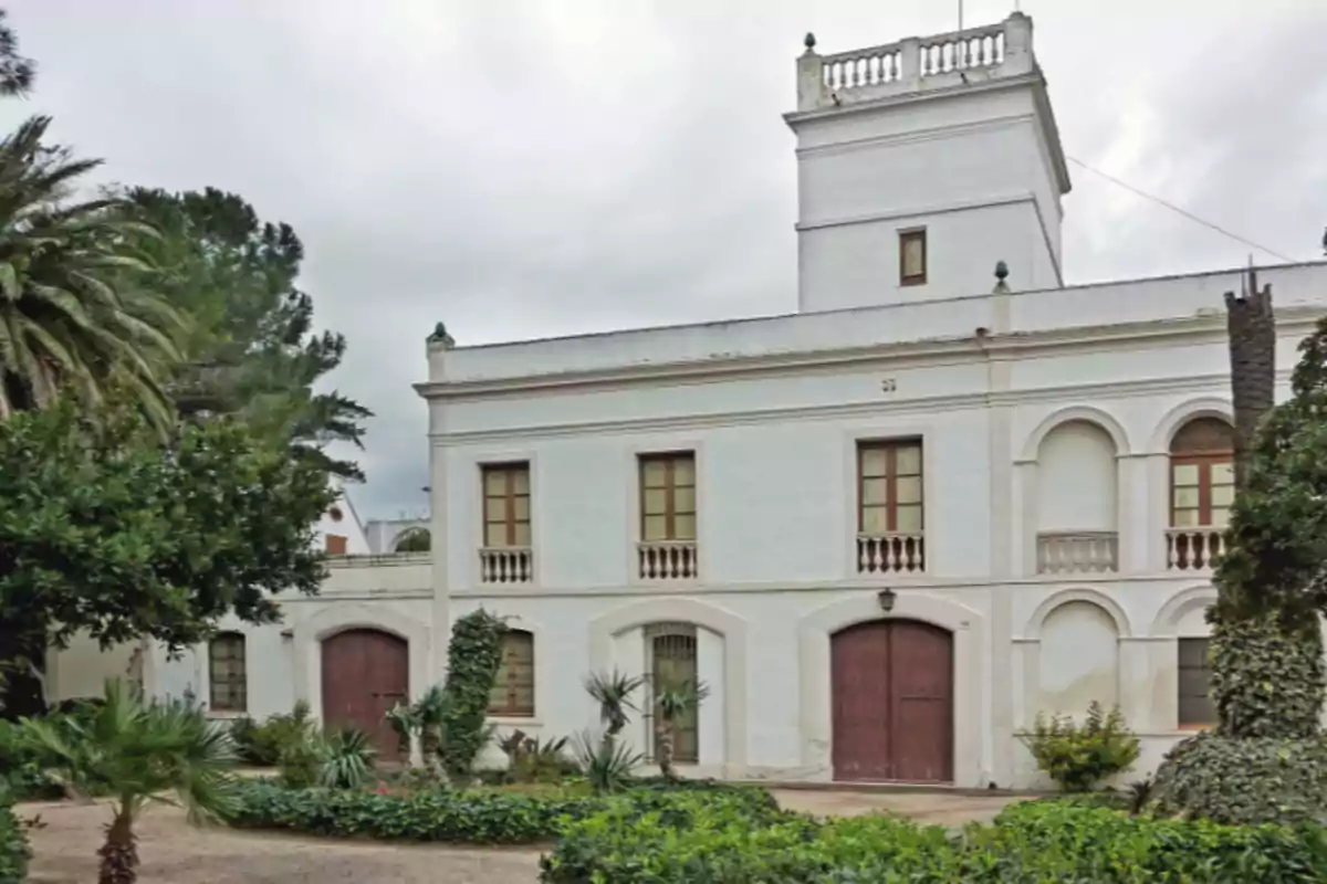 Fachada de una casa antigua de dos pisos pintada de blanco con puertas y ventanas de madera rodeada de vegetación y árboles Fachada de una casa antigua de dos pisos pintada de blanco con puertas y ventanas de madera rodeada de vegetación y árboles
