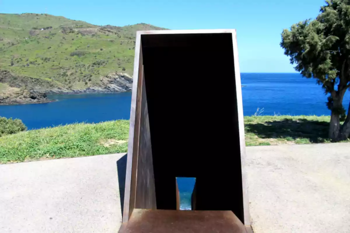 Escultura de metall amb forma rectangular situada davant d’un paisatge de turons verds i mar blau sota un cel serè Escultura de metall amb forma rectangular situada davant d’un paisatge de turons verds i mar blau sota un cel serè