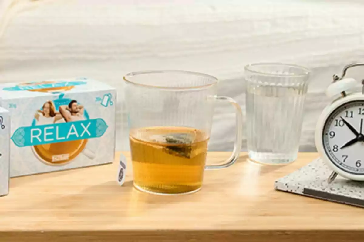 Tassa de te al costat d’un got d’aigua, una capsa d’infusions i un rellotge despertador sobre una taula de fusta Tassa de te al costat d’un got d’aigua, una capsa d’infusions i un rellotge despertador sobre una taula de fusta