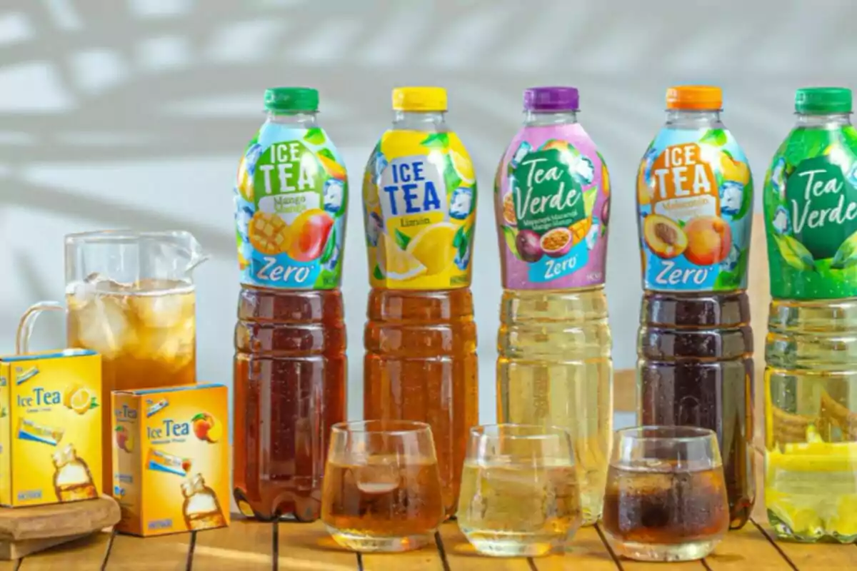 Botellas de té helado de diferentes sabores junto a vasos con hielo, una jarra y cajas de té sobre una mesa de madera Botellas de té helado de diferentes sabores junto a vasos con hielo, una jarra y cajas de té sobre una mesa de madera