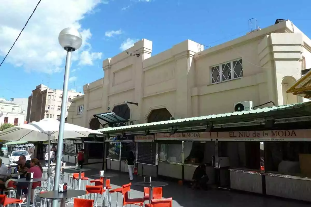 Persones assegudes en una terrassa amb cadires vermelles davant d’un edifici de mercat de color clar en un dia assolellat Persones assegudes en una terrassa amb cadires vermelles davant d’un edifici de mercat de color clar en un dia assolellat