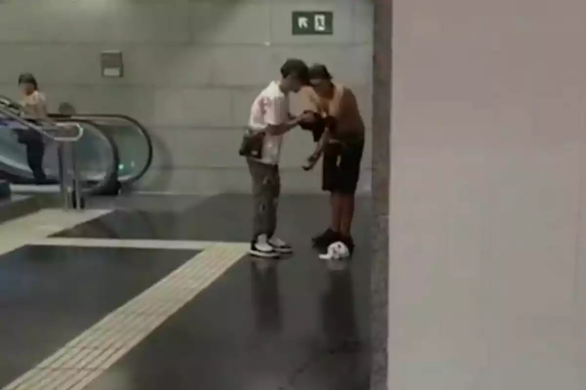 Dos personas conversan junto a una pared en una estación de metro mientras una pelota de fútbol está en el suelo cerca de ellos y al fondo una mujer sube por una escalera eléctrica. Dos personas conversan junto a una pared en una estación de metro mientras una pelota de fútbol está en el suelo cerca de ellos y al fondo una mujer sube por una escalera eléctrica.