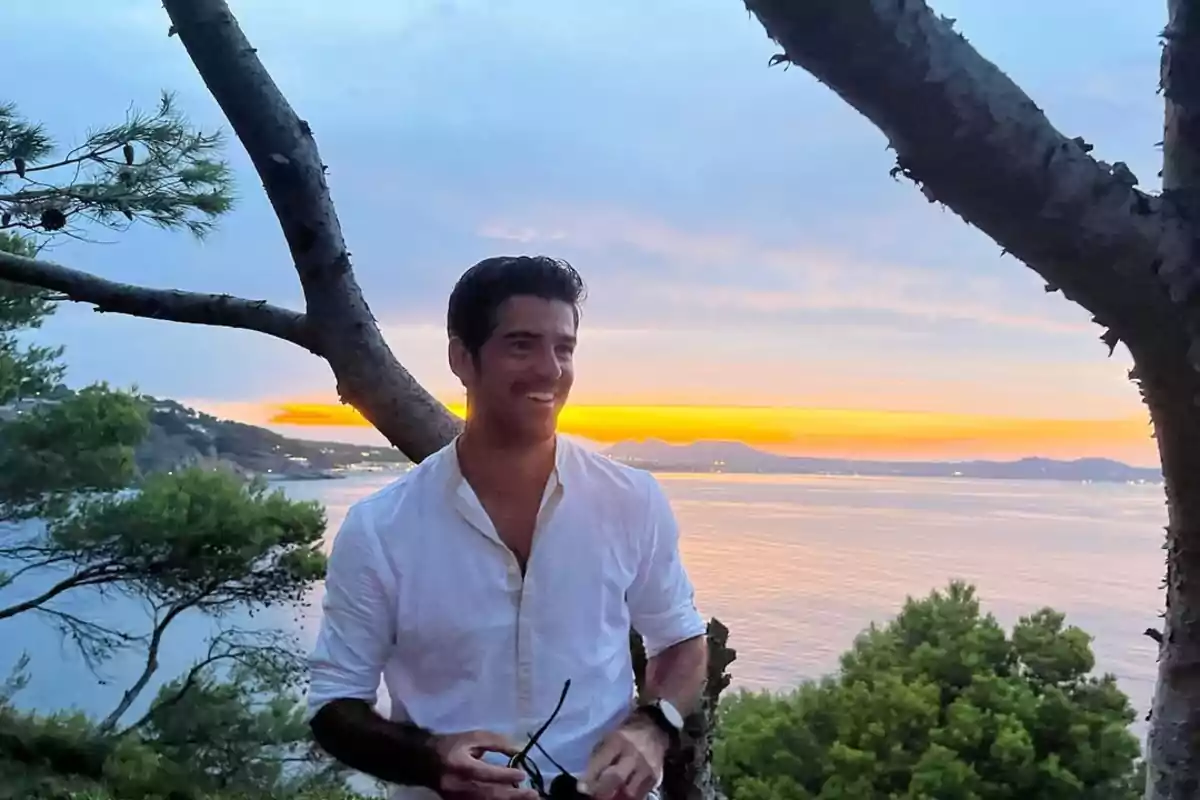 Home somrient amb camisa blanca dret entre arbres davant del mar al capvespre Home somrient amb camisa blanca dret entre arbres davant del mar al capvespre