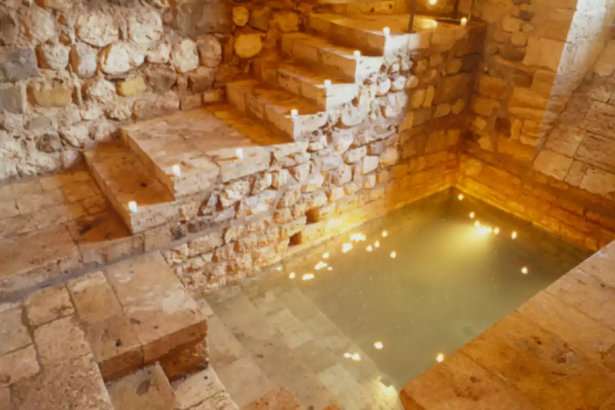 Escala de pedra antiga que descende cap a una piscina d’aigua poc profunda envoltada de murs de pedra il·luminada per petites llums flotants. Escala de pedra antiga que descende cap a una piscina d’aigua poc profunda envoltada de murs de pedra il·luminada per petites llums flotants.