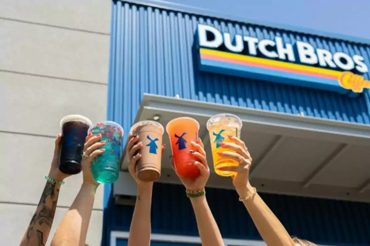Varias manos levantan vasos de bebidas coloridas frente a una sucursal de Dutch Bros Varias manos levantan vasos de bebidas coloridas frente a una sucursal de Dutch Bros