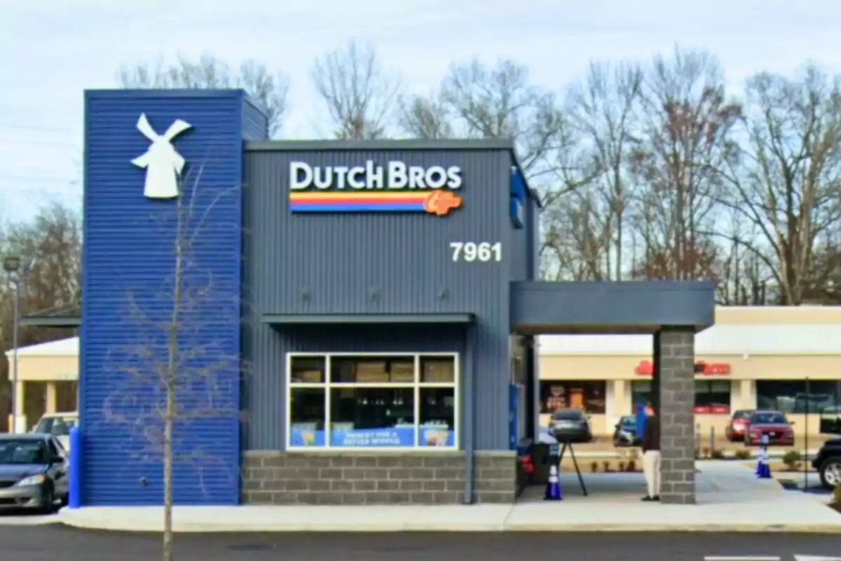 Sucursal de Dutch Bros Coffee con fachada azul y gris, un molino de viento como logotipo y un número 7961 en la pared, ubicada en una zona comercial con autos estacionados y árboles sin hojas al fondo Sucursal de Dutch Bros Coffee con fachada azul y gris, un molino de viento como logotipo y un número 7961 en la pared, ubicada en una zona comercial con autos estacionados y árboles sin hojas al fondo