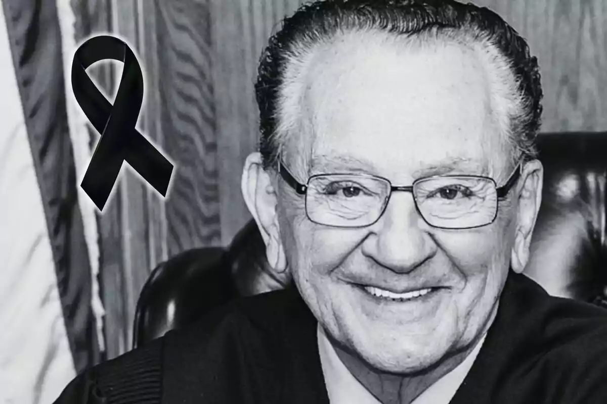 Hombre mayor con gafas sonriendo en una fotografía en blanco y negro con un moño negro de luto en la esquina izquierda