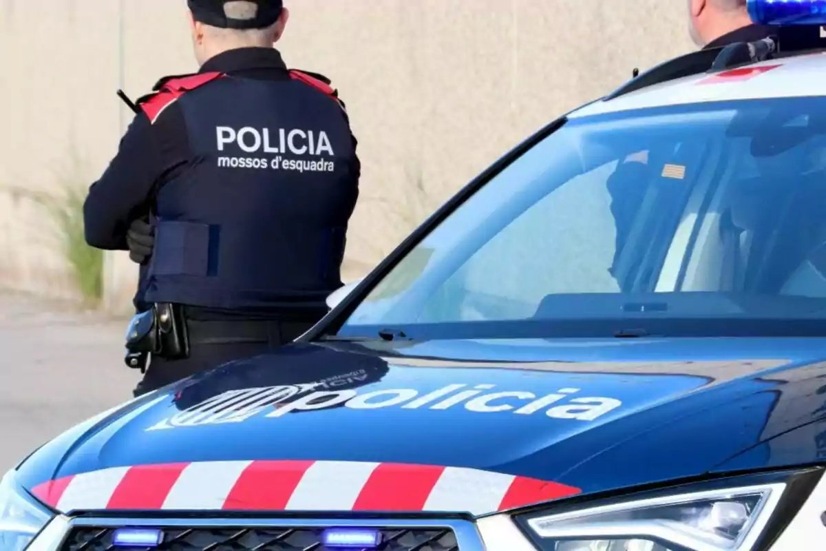 Agent dels Mossos d'Esquadra d'esquena al costat d'un cotxe patrulla blau amb els llums encesos