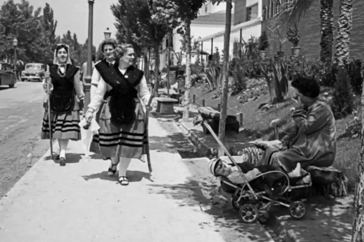 Tres mujeres caminan por una acera vestidas con trajes tradicionales mientras una mujer sentada cuida a un niño en un cochecito antiguo Tres mujeres caminan por una acera vestidas con trajes tradicionales mientras una mujer sentada cuida a un niño en un cochecito antiguo