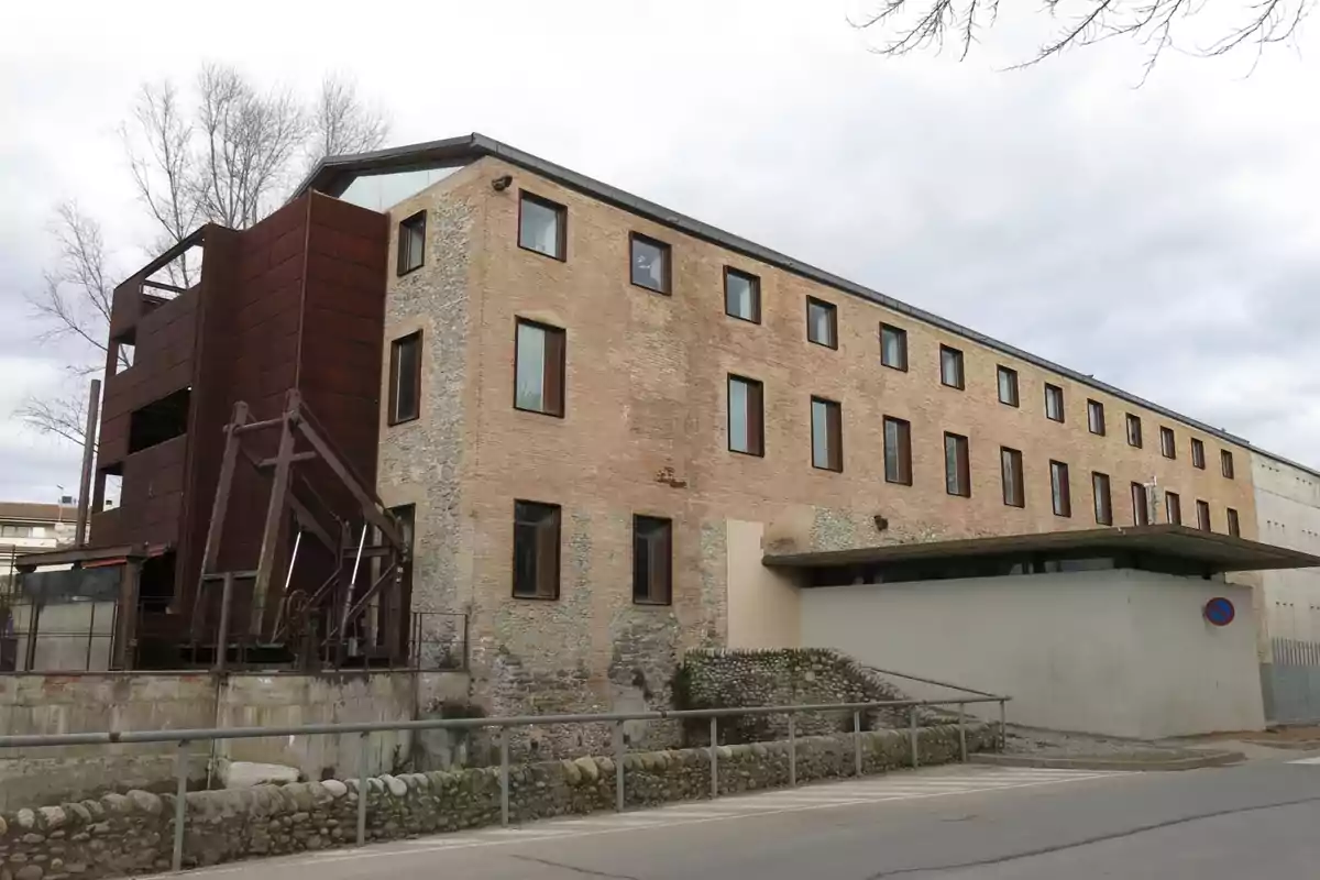 Edificio de ladrillo y piedra con varias ventanas rectangulares y una estructura metálica en uno de sus extremos junto a una calle y un muro bajo de piedras Edificio de ladrillo y piedra con varias ventanas rectangulares y una estructura metálica en uno de sus extremos junto a una calle y un muro bajo de piedras