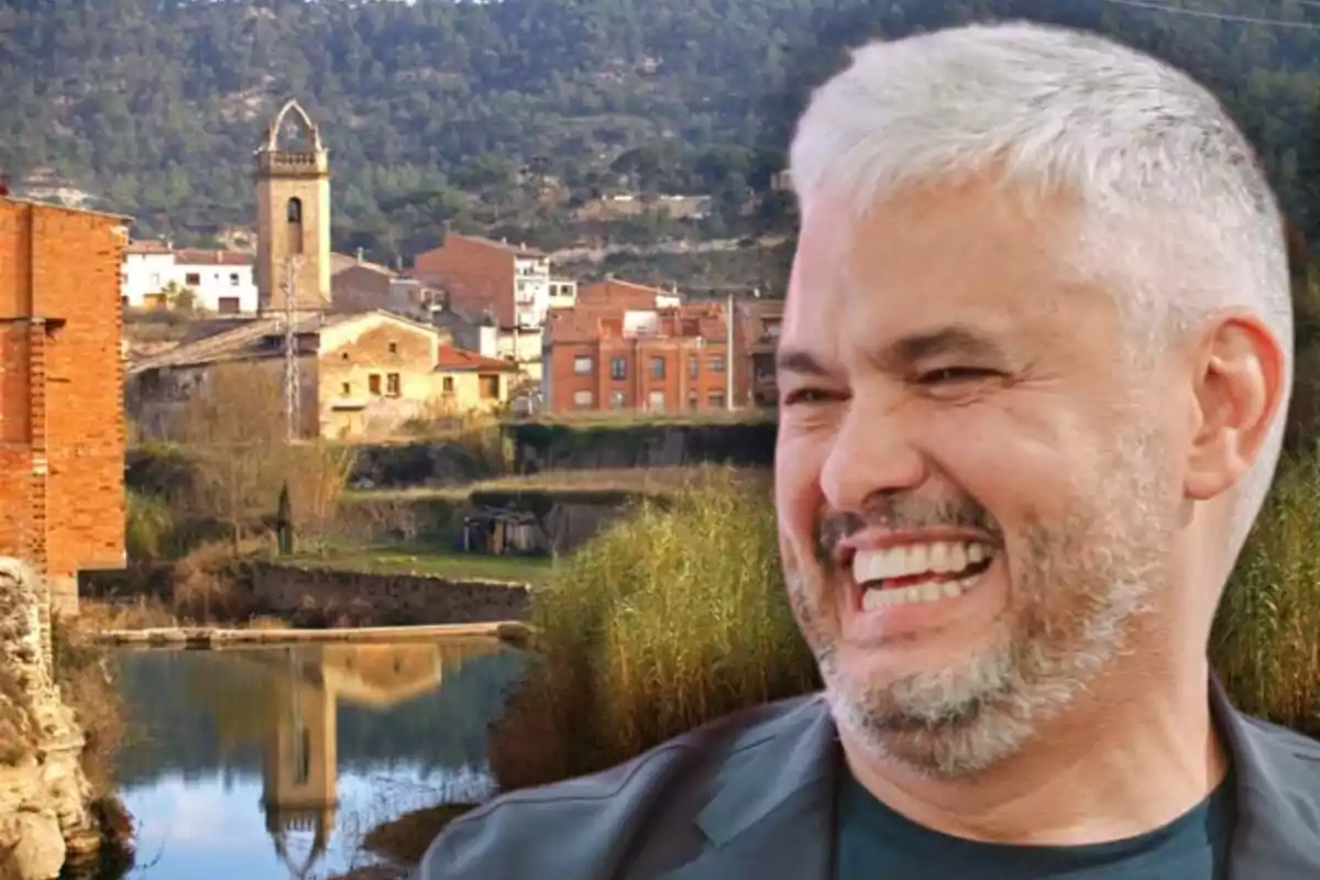 Home de cabells canosos i barba somrient en primer pla amb un poble i un campanar reflectits a l’aigua al fons