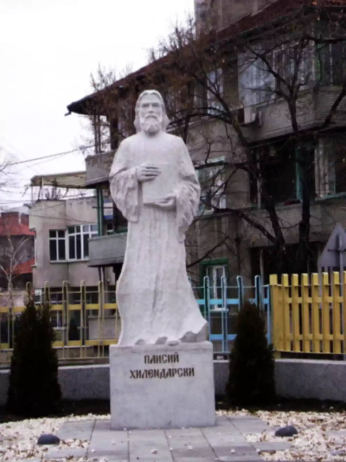 Estàtua de Paisii Hilendarski sostenint un llibre amb totes dues mans en un entorn urbà Estàtua de Paisii Hilendarski sostenint un llibre amb totes dues mans en un entorn urbà