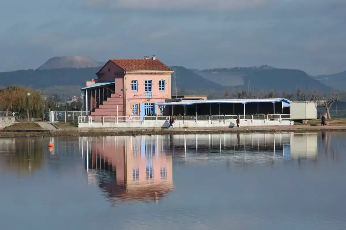 Edifici rosa amb finestres blaves reflectit en un llac tranquil amb muntanyes al fons i cel parcialment ennuvolat Edifici rosa amb finestres blaves reflectit en un llac tranquil amb muntanyes al fons i cel parcialment ennuvolat