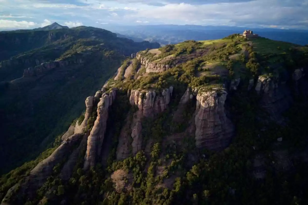 Vista aèria d’una muntanya amb formacions rocoses i vegetació, al cim s’observa una edificació aïllada sota un cel parcialment ennuvolat. Vista aèria d’una muntanya amb formacions rocoses i vegetació, al cim s’observa una edificació aïllada sota un cel parcialment ennuvolat.