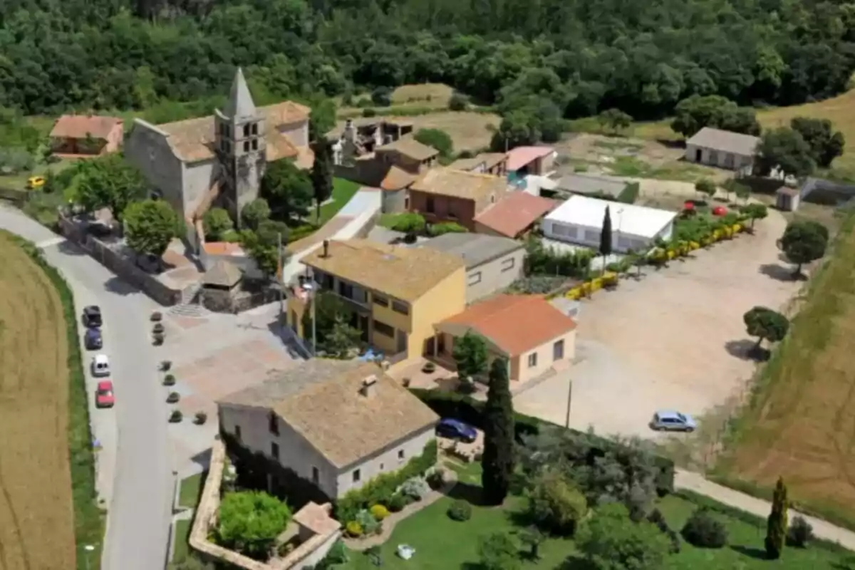 Vista aérea de un pequeño pueblo rural con varias casas de tejado rojo, una iglesia con campanario y áreas verdes rodeadas de campos y árboles Vista aérea de un pequeño pueblo rural con varias casas de tejado rojo, una iglesia con campanario y áreas verdes rodeadas de campos y árboles