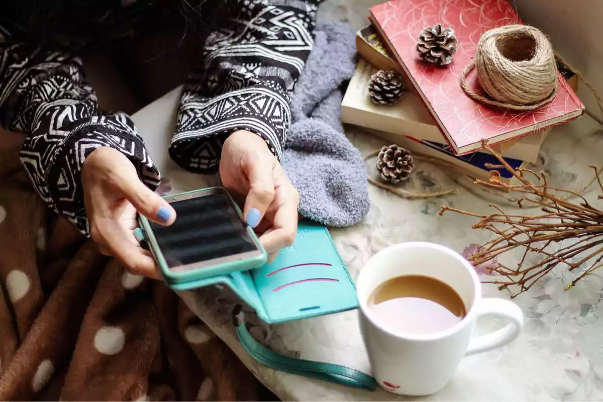 Persona amb jersei de patrons geomètrics utilitzant un telèfon mòbil al costat d’una tassa de cafè, llibres, pinyes seques, fil i branques sobre una taula