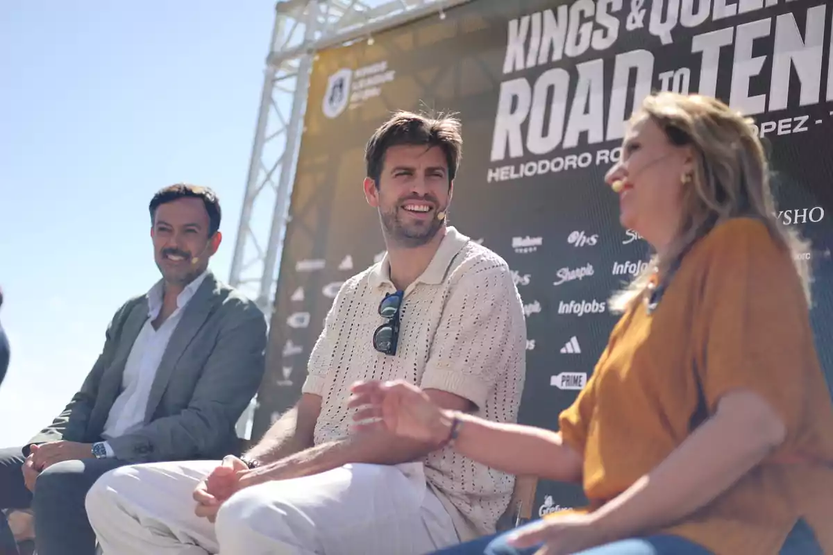 Tres persones assegudes conversant i somrient en un esdeveniment a l’aire lliure amb un cartell de fons que diu Kings and Queens Road to Ten Tres persones assegudes conversant i somrient en un esdeveniment a l’aire lliure amb un cartell de fons que diu Kings and Queens Road to Ten