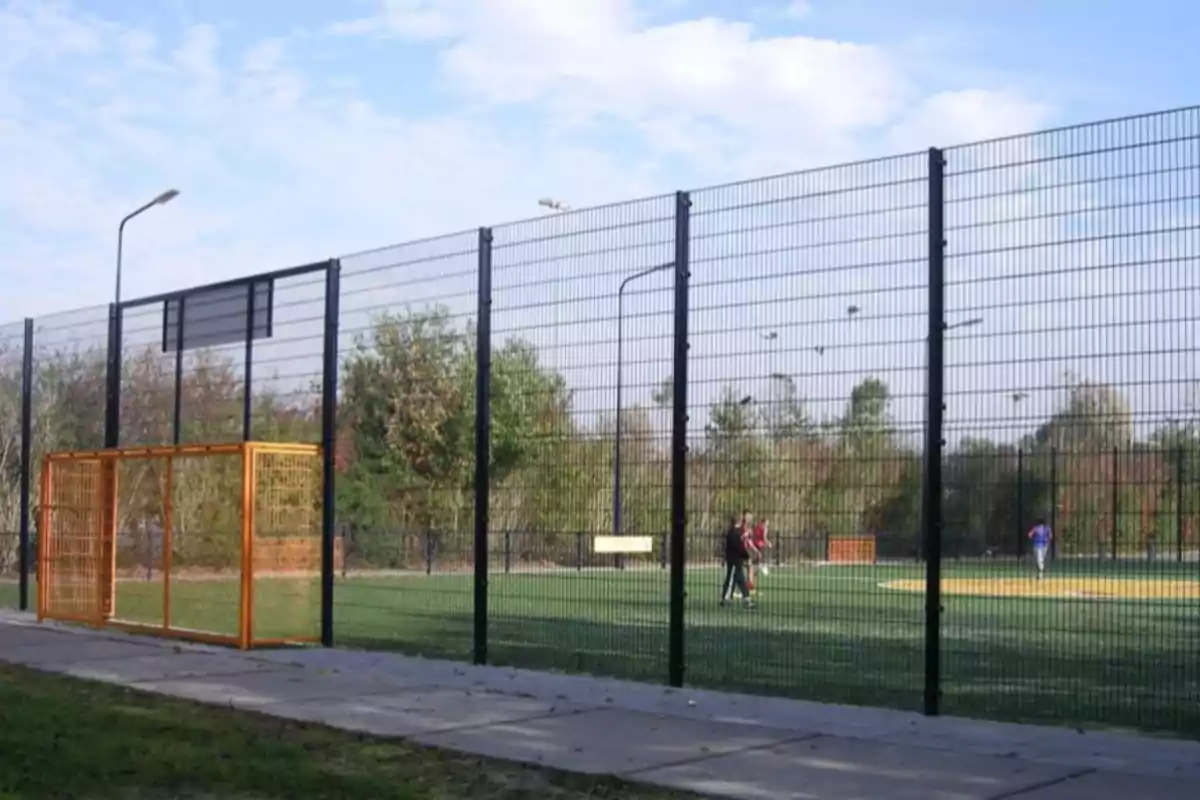 Pista esportiva a l’aire lliure envoltada per una tanca alta, amb algunes persones jugant a l’interior i arbres al fons. Pista esportiva a l’aire lliure envoltada per una tanca alta, amb algunes persones jugant a l’interior i arbres al fons.