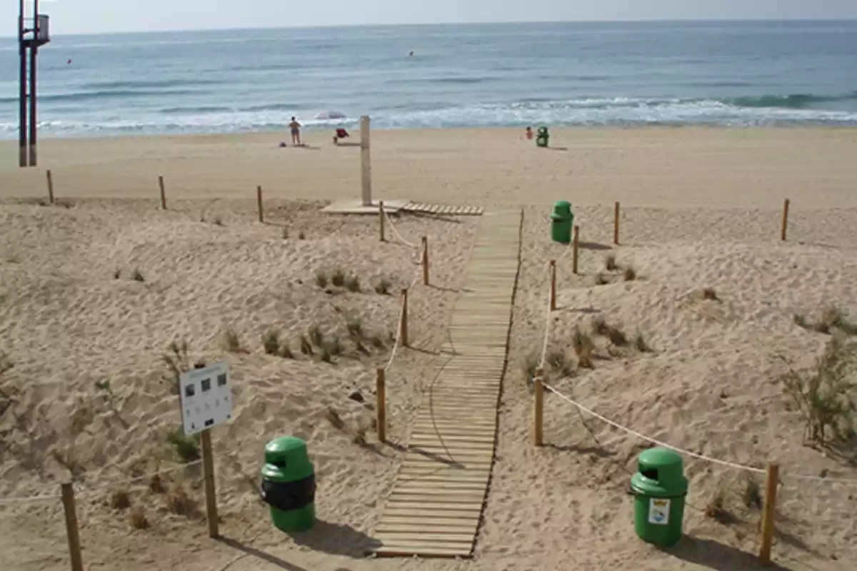 Pasarela de madera sobre la arena que conduce a la orilla del mar con papeleras verdes y algunas personas disfrutando de la playa Pasarela de madera sobre la arena que conduce a la orilla del mar con papeleras verdes y algunas personas disfrutando de la playa