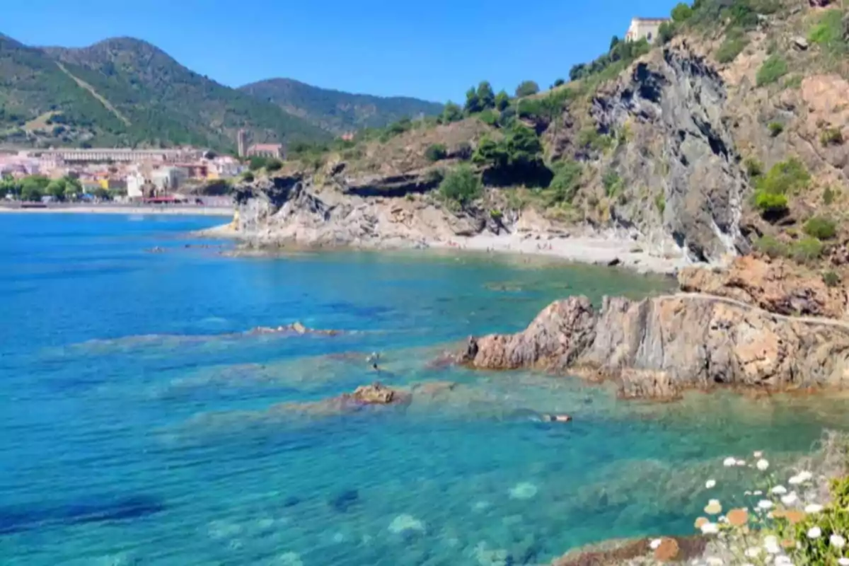Platja d’aigües cristal·lines envoltada de roques i vegetació, amb muntanyes al fons i un poble costaner a l’esquerra. Platja d’aigües cristal·lines envoltada de roques i vegetació, amb muntanyes al fons i un poble costaner a l’esquerra.